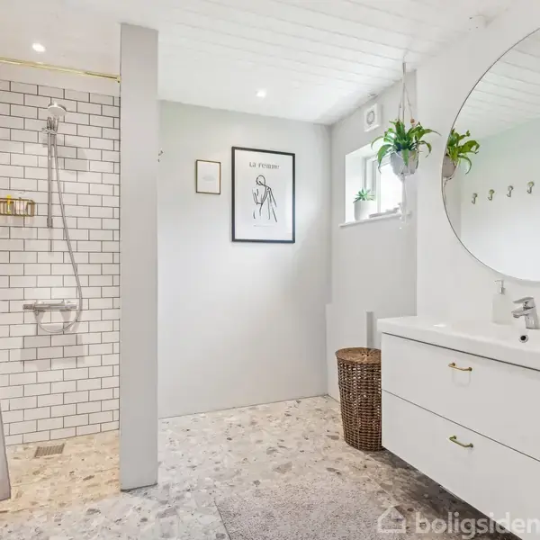 En moderne brusekabine med hvide fliser og en grå forhæng står i en lys badeværelseskontekst. En rund spejl hænger over en hvid vask, ved siden af en plante og en plakat.