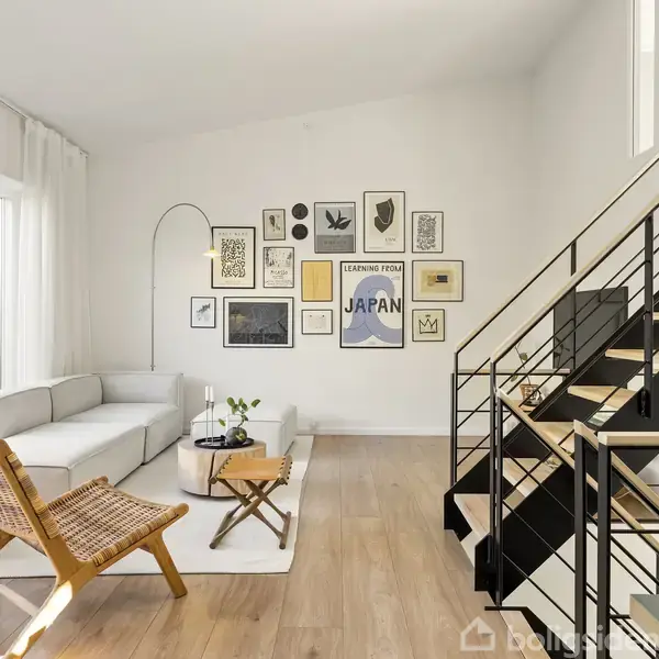 Sofagruppe med gråt hjørnesofa og træstol, placeret i stue med kunstnerisk vægdekoration af forskellige billeder. Moderne trappe til højre og stort vindue til venstre med lukkede gardiner.
