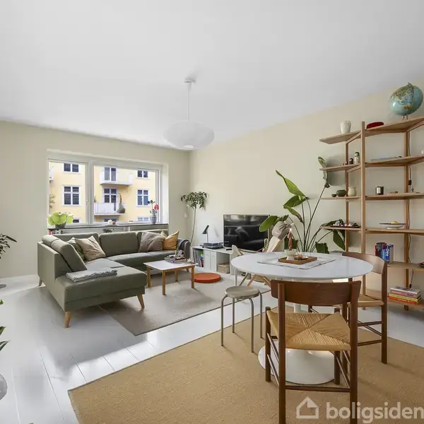 To stole står på en lille altan med blomster i potter i midten. Altanen vender mod en gul bygning med altaner. Indendørs er der store grønne planter.