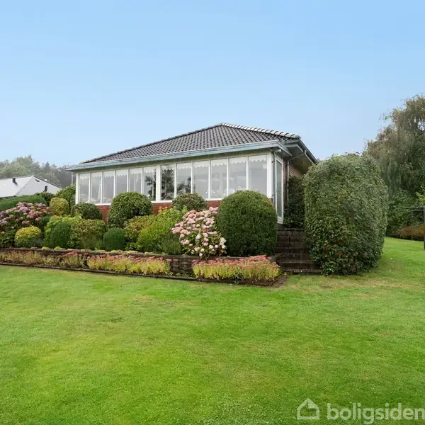 Et enfamiliehus med store vinduer omgivet af velplejet have med buske og blomsterbede. Huset står på en pæn græsplæne i et grønt område med andre huse i baggrunden.