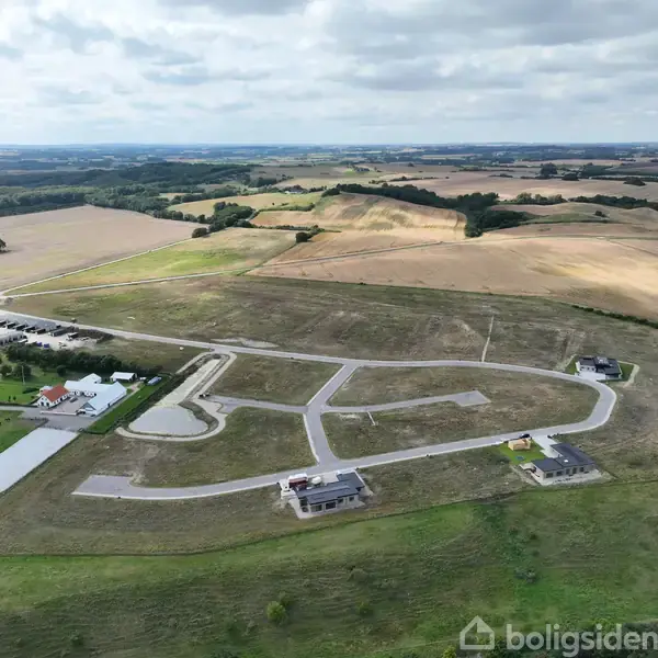 Bygninger placeret langs snoede veje i et landskab med store græsarealer og åbne marker under en skyet himmel. En parkeringsplads og nogle landlige bygninger ses i baggrunden.