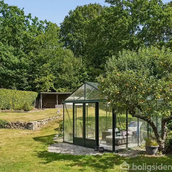 Rødstenshus med rødt tegltag står i en have med flagstang og hævet terrasse under en parasol. Græsplæne og træer omgiver huset under en blå himmel med skyer.