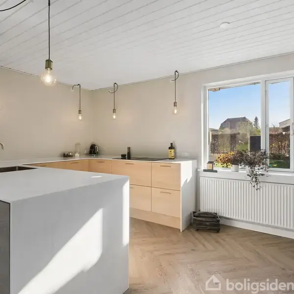 Et moderne køkken med lyst trægulv og lyse træskabe. Loftslamper hænger ned fra det hvide loft. Et vindue med udsigt til haven. Moderne bordplade med indbygget vask.