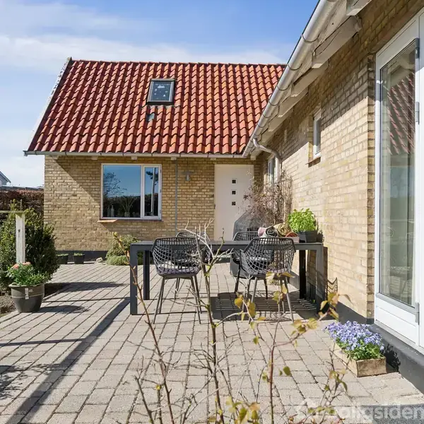 En murstensvilla med rødt tegltag har en flisebelagt terrasse med sorte havemøbler og krukker med planter. Villaen ligger i et solrigt område med en velplejet have.