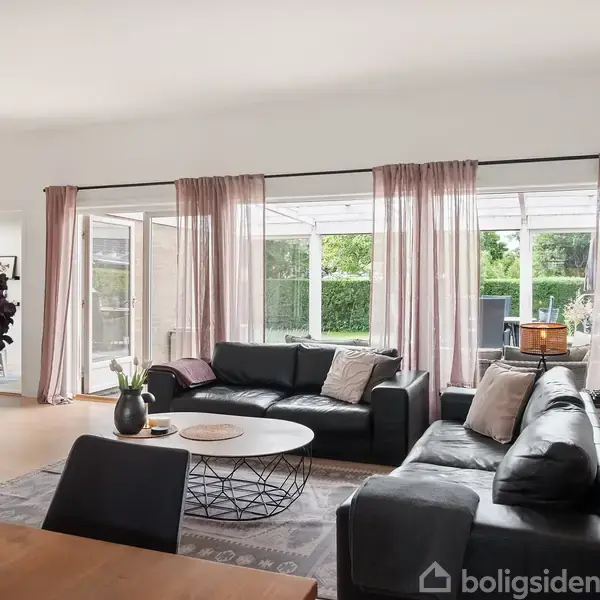 Sort lædersofa står i en moderne stue, omgivet af lyserøde gardiner og glasdøre, der åbner ud mod en grøn baghave. Pejse- og kafédekorationer skaber en hyggelig atmosfære.