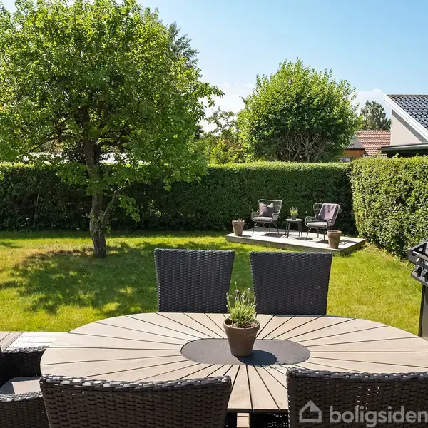 Et rundt havebord med seks stole står på en terrasse. En grill er ved siden af. I baggrunden er der to stole og en sofa i en indhegnet have med græsplæne og træer.