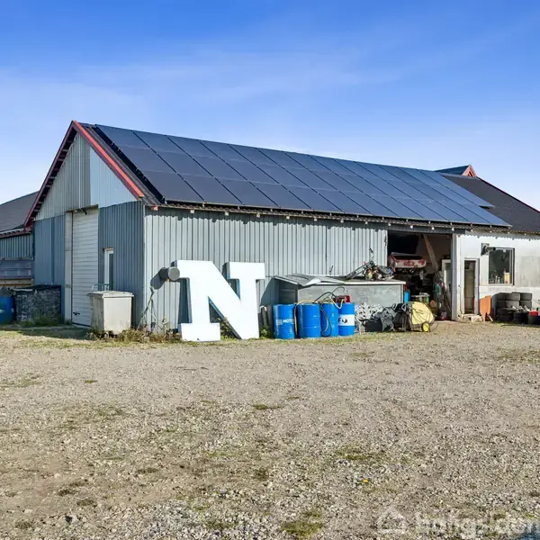 En grå bygning med tagdækkede solpaneler ses på en grusbelagt grund. Foran bygningen står et stort, hvidt bogstav "N" samt tønder og en vintage bil.
