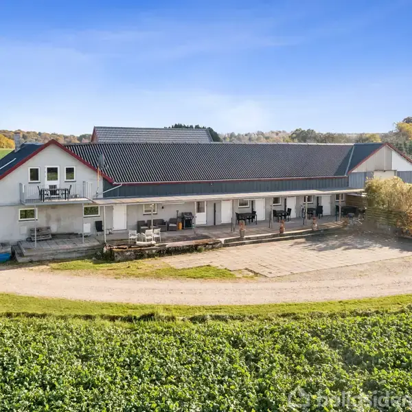 En lang, hvid bygning med mørkt skråt tag står på en grusvej, omgivet af marker. Flere vinduer og altaner ses på bygningen, og campingvogne er placeret til højre.