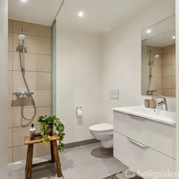 Brusebad med glasvæg, beige fliser og brun badehåndklæde. Håndvask med spejl til højre. Skammel med plante står midt. Vaskemaskine og tørretumbler i forgrunden til højre.