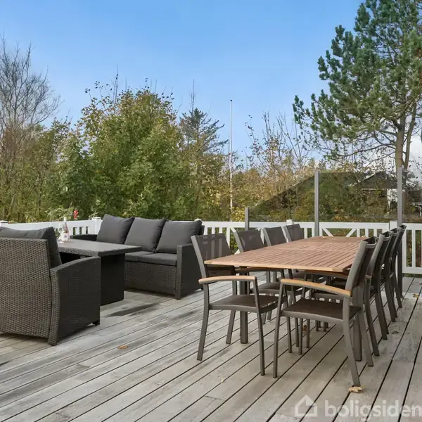 Terrassemøbler står på et trædæk med sofa og spisebord med stole. Omkring terrassen er der et hegn og træer, hvilket giver en hyggelig, naturlig atmosfære.