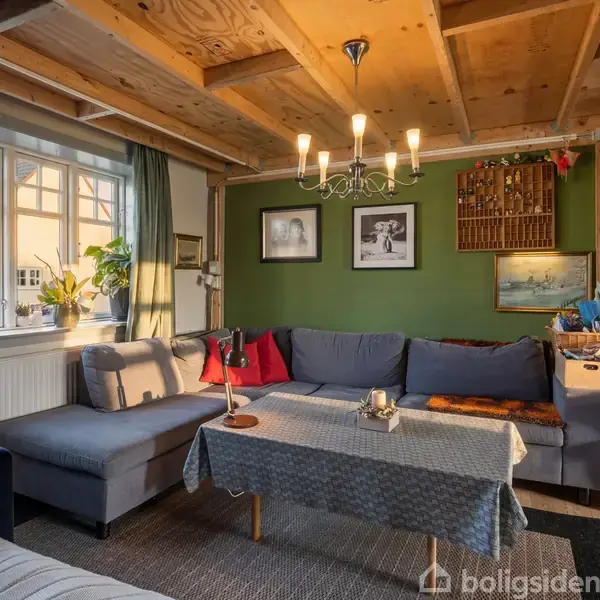 En stue med en grå hjørnesofa centreret omkring et bord med dug. Der hænger en lysekrone i det træbeklædte loft. Vindueskarm med planter og farvede vaser.