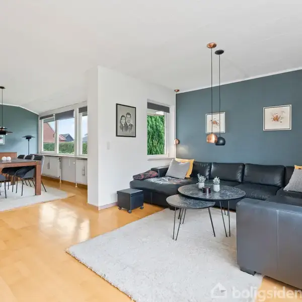 En moderne stue med en sort hjørnesofa og runde sofaborde på et lyst tæppe. Ved siden af er en spiseplads med et bord og stole. Rummet har lyse trægulve og store vinduer.