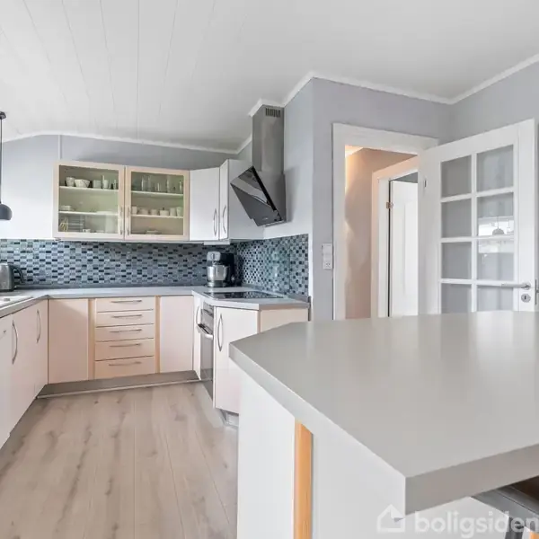 Køkken med hvide skabe og glaslåger. Der står en kaffemaskine og brødrister på bordet. Rummet har lysegrå vægge, trælaminatgulv, og et flisebelagt komfurhjørne.