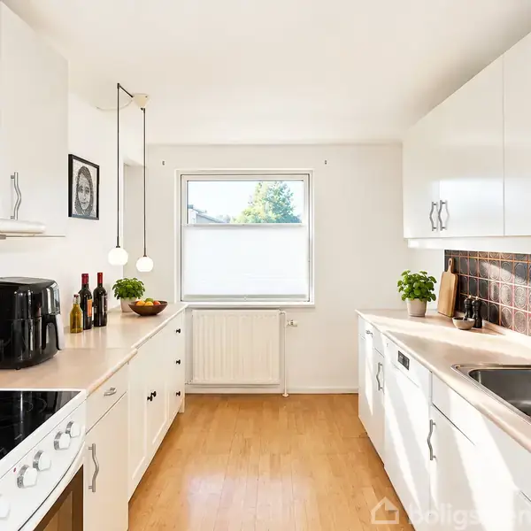 Et moderne køkken med hvide skabsfronter og en sort kogeplade. En airfryer og vinflasker står på bordet, mens vinduet giver naturligt lys. Planter tilføjer grønne nuancer.