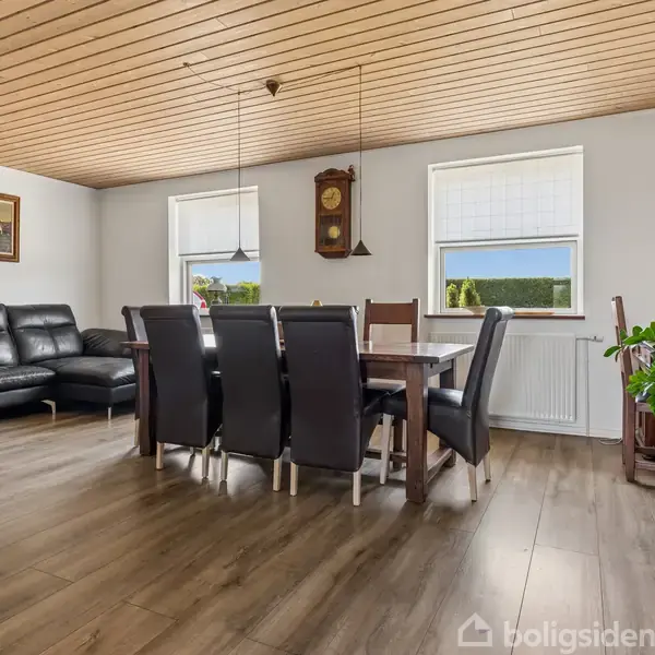 Spisebord med otte stole står i en stue ved siden af en sort lædersofa. Der hænger en pendellampe over bordet, og en vægklocke er placeret mellem vinduerne.