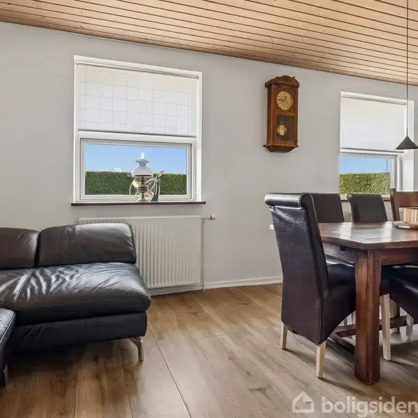 Sort læderhjørnesofa står langs væggen. Spisebord med seks stole er placeret ved vinduerne i lyse rum med trægulv og træloft. Vægur hænger mellem vinduerne.