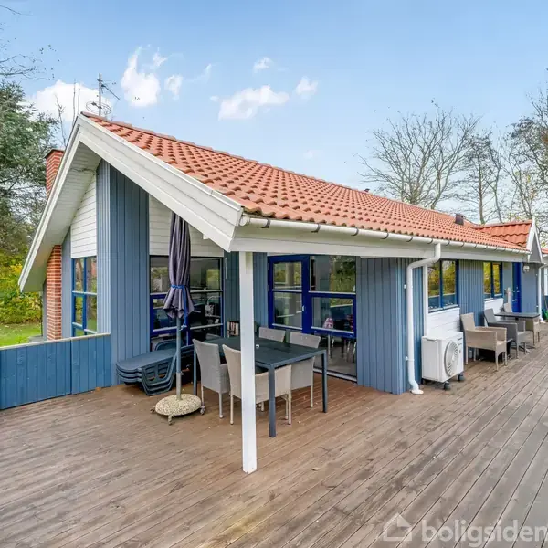 Et blåmalet hus med rødt tag står på en træterrasse. Der er et udendørs spisebord med stole. Omkring huset er der træer og grønne omgivelser.