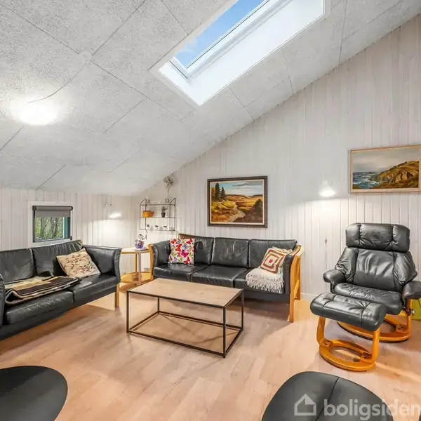 Stue med sorte lædersofaer og lænestol, står omkring et sofabord. Rummet er lyst med ovenlys og vægkunst. Trægulv og hvide vægge skaber en moderne og hyggelig atmosfære.
