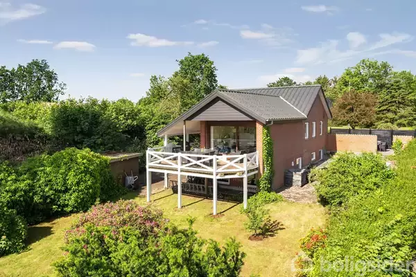 Rødt murstenshus med hævet terrasse, omgivet af frodig have med buske og træer. Solrig dag med blå himmel og få skyer. Ingen tekst er synlig i billedet.