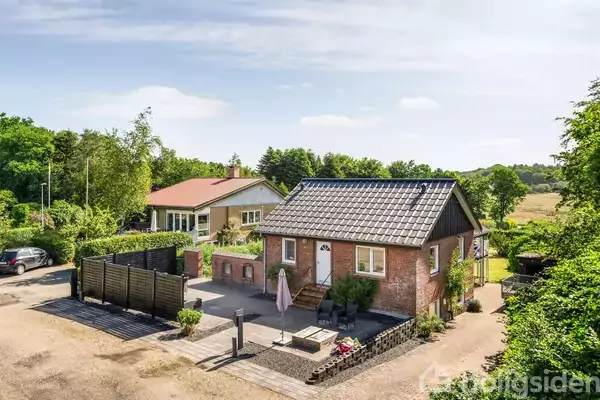Et rødt murstenshus med sort tag står i en have omkranset af grøn vegetation. Foran huset er der en terrasse med parasol, omgivet af hække. Vandret sollys skaber en lys atmosfære.