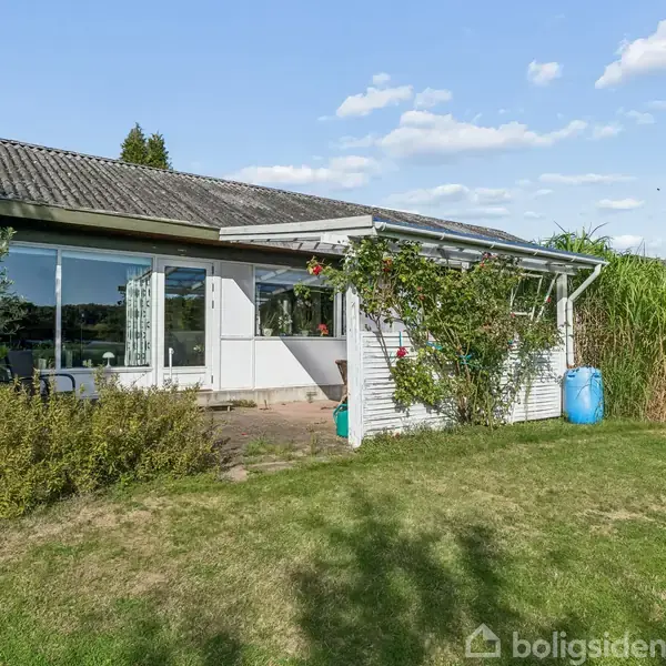 Et hus med store vinduer og en åben terrasse er omkranset af en grøn have med buske og træer under en blå himmel.