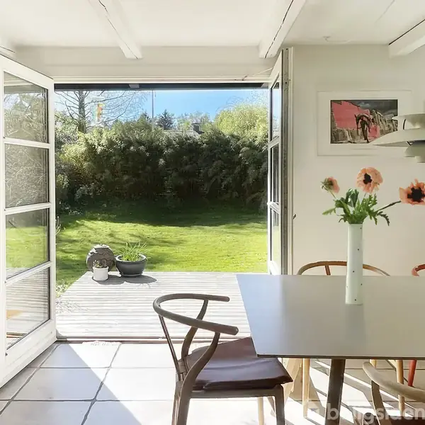 Et spisebord med en vase med røde tulipaner står i et lyst rum. Døre er åbne mod en terrasse og en have med græs og buske.