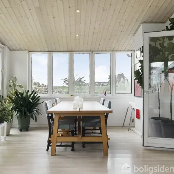 Et lyst rum med træloft indeholder et langt bord omgivet af stole. Der er grønne planter ved vinduerne, og en dør står åben til en anden del af huset.