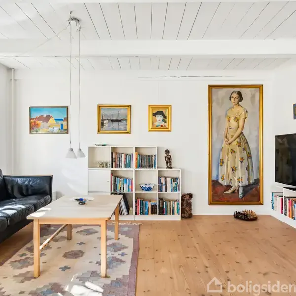 Sort lædersofa ved træbord i stue med hvide vægge og loft. Bøger på hylder, stor maleri af kvinde, og et tv ved siden af i hyggeligt, indrettet rum.