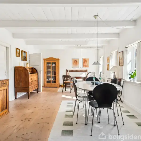 Et spisebord med sorte stole står i et lyst rum. Bordet er dækket med lamper over. Til højre er store vinduer, til venstre en brændeovn og malerier.