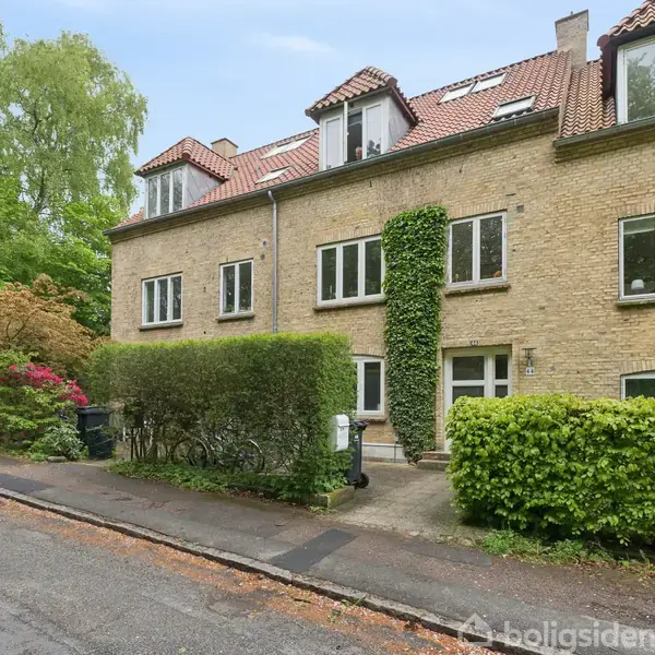 Rækkehus i gule mursten med rødt tegltag placeret i en grøn, frodig boligområde. Der er hække og træer langs fortovet, som skaber en indbydende atmosfære.