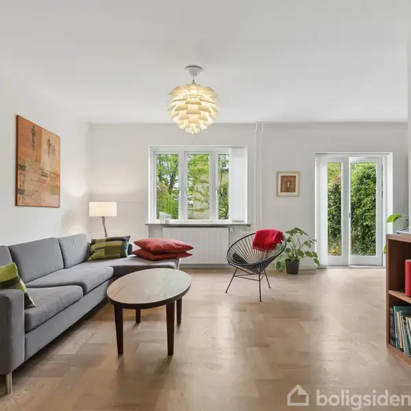 Grå sofa med farvede puder står i et lyst rum med trægulv. Ved siden af er et rundt bord og en moderne stol. Vinduer åbner ud til en have, mens indretningen omfatter planter og kunst på væggene.
