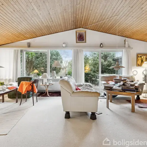 Stue med hvide vægge og trægulvloft, indeholder to grønne sofaer, en hvid lænestol og diverse borde med bøger og lamper. Store vinduer viser grønne buske udenfor.