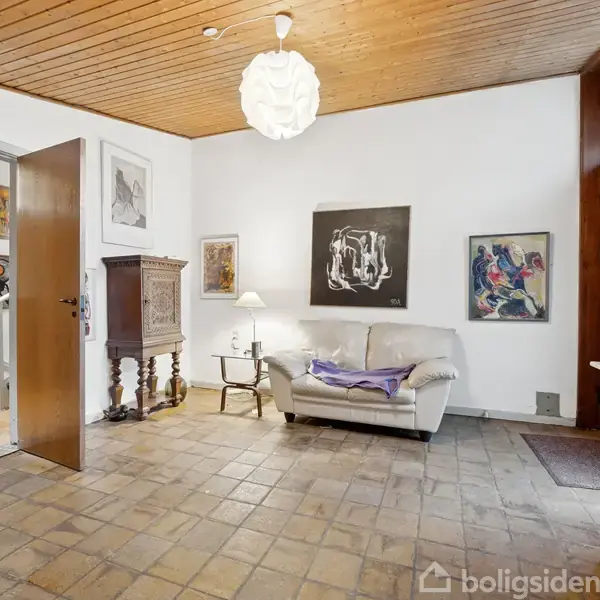 En stue med en beige sofa der er dekoreret med puder, omgivet af kunstmalerier på væggene. Fliser på gulvet, træloft, og en åben dør viser en anden del af huset.