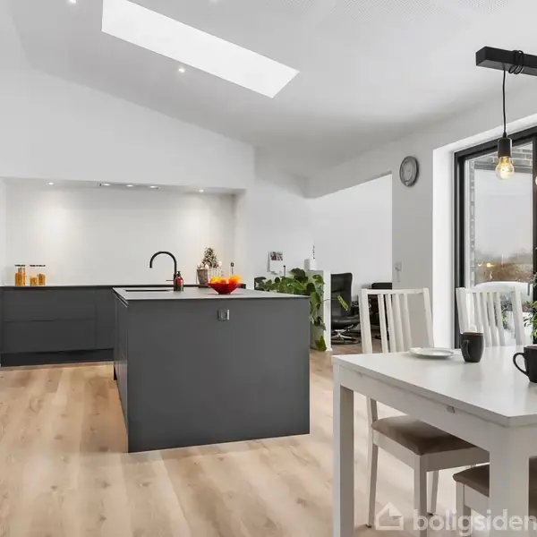 Køkken-alrum med moderne, mørkegrå køkkenø, omgivet af hvide vægge og trælaminatgulv. Spisebord med hvide stole står ved store vinduer. Loftslamper hænger over bordet. Ingen tekst synlig.