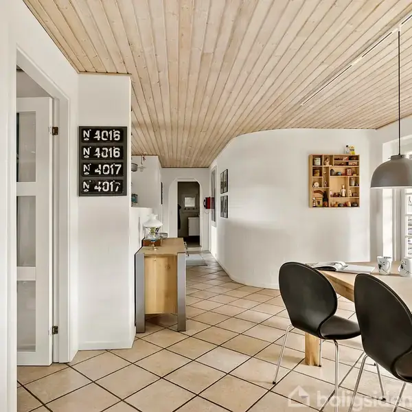 Moderne køkkenalrum med spisebord. Sorte stole står omkring bordet under to pendellamper. Lyse klinkegulve og træloft. Vægpynt inkluderer hylder og skilte med "N 4016", "N 3016", "N 4017".