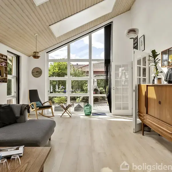 Stue med grå sofa og træskænk. Store vinduer lukker lys ind, og skydedøre leder ud til en terrasse med planter. Loftet har synlige bjælker og en moderne indretning.