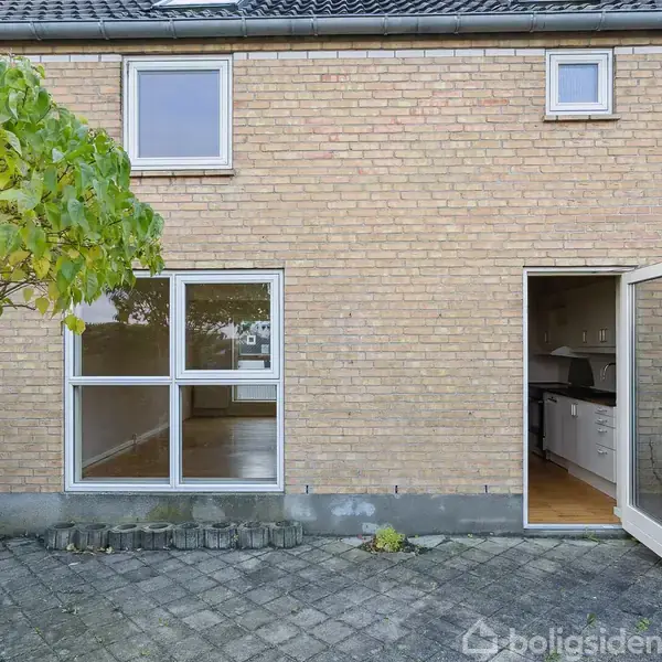 Et murstenshus med to vinduer og en åben dør. Døren fører til et køkken. En flisebelagt terrasse med et træ og grønne buske i en have.