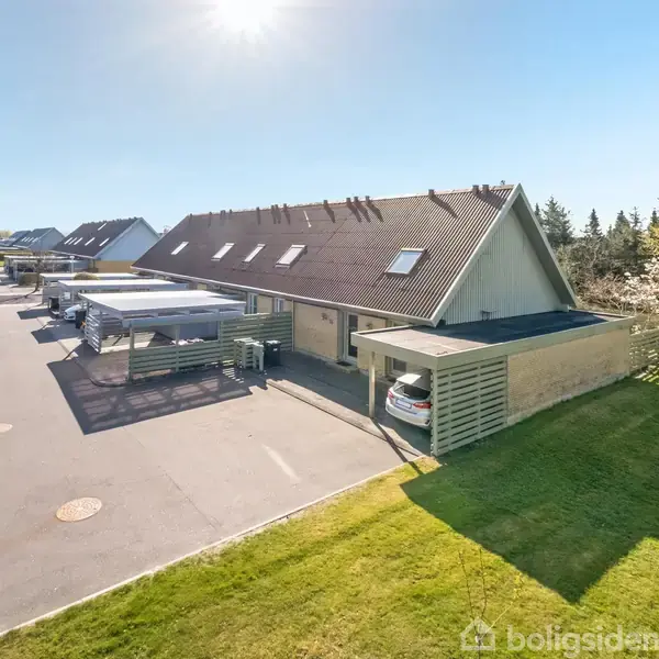 Rækkehus med skråt tag står stille i solrigt vejr. En bil er parkeret under carporten ved siden af huset. Omgivet af grønne græsplæner og en række af lignende huse.