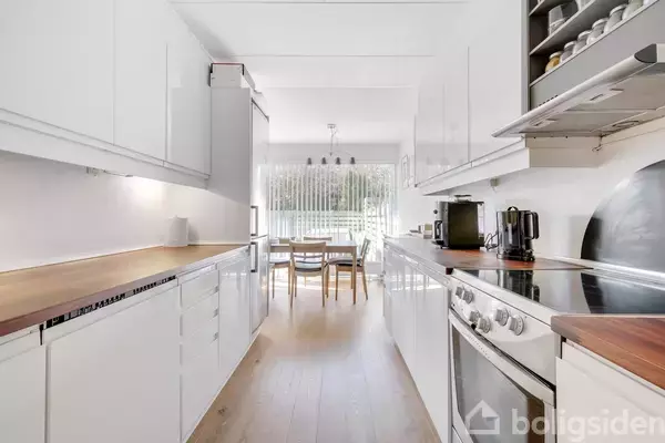 Et moderne køkken med hvide skabe og træbordplader, indeholder en ovn og kaffemaskine. I baggrunden ses et spisebord med stole ved et stort vindue, der slipper lys ind.