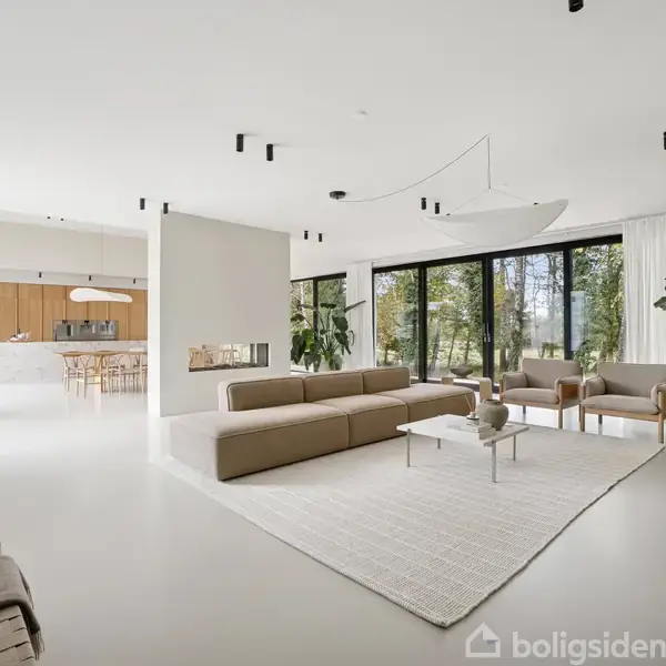 En lys stue med beige sofaer og et lyst tæppe midt på gulvet, omgivet af store, grønne planter. I baggrunden ses et åbent køkken med træmøbler og store vinduer med udsigt til naturen.