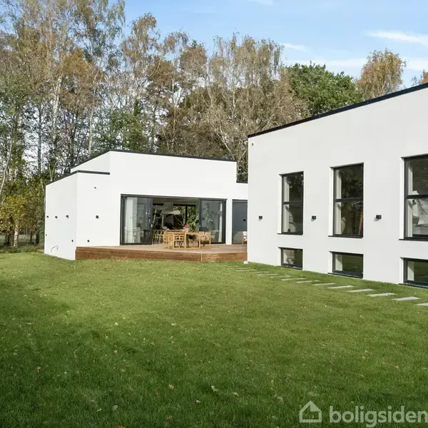 En moderne hvid bygning med store vinduer står stille i en velplejet have med grøn græsplæne. Omkring bygningen er der skovbevoksning under en blå himmel.
