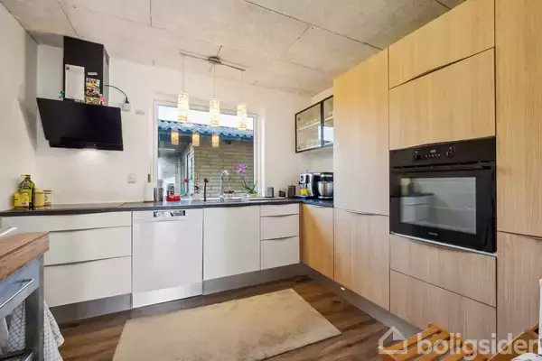 Køkken med moderne apparater, herunder ovn og opvaskemaskine, opstillt i et hjørne. Et vindue ved vasken slipper naturligt lys ind. Lyse træskabe omgiver rummet, der har trægulve.