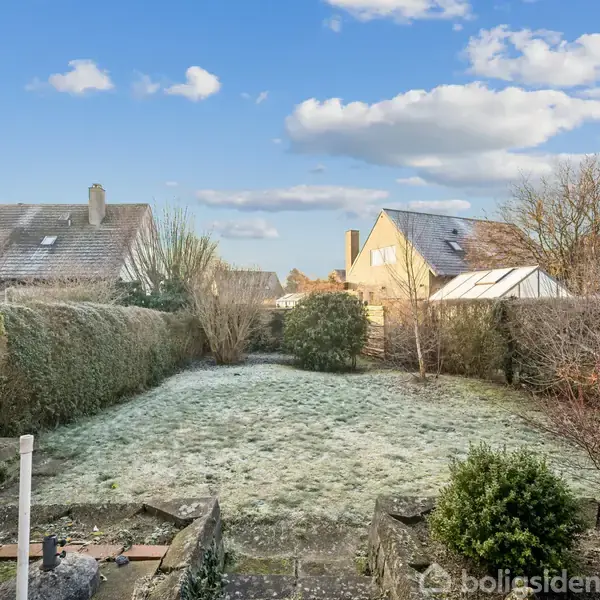 En frostbelagt baghave er omkranset af tætte buske og hegn, med udsigt til naboernes hustage under en klar blå himmel med spredte skyer.