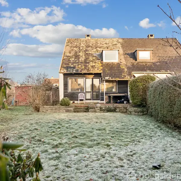Et hus med skråtag står i en have med rimdækket græs. Buske omkranser området, mens blå himmel og skyer ses i baggrunden.