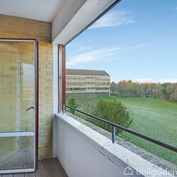 En altan med glasdør, der åbner mod en stor grøn græsplæne. I baggrunden ses en murstensbygning omgivet af træer under en klar blå himmel.