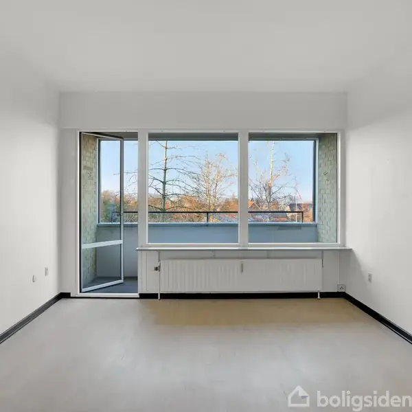 Et tomt rum med lys beige gulv og hvide vægge, store vinduer og en glasdør, der åbner til en balkon. Udsigt til træer udenfor i dagslys.