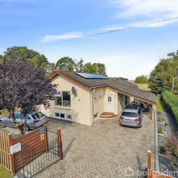 Hus med solpaneler på taget, to parkerede biler i indkørslen, omgivet af en have med træer og en hæk. En låge markerer indgangen til ejendommen.