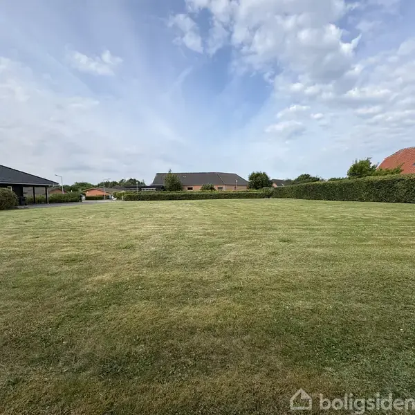 En stor græsplæne strækker sig ud i en have omkranset af lave hække. I baggrunden ses flere huse med tage, og himlen er delvist overskyet.