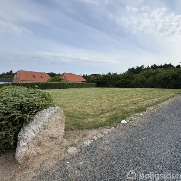 Græsplæne omgivet af hække og buske. To huse med røde tage ses i baggrunden, mens en grussti fører til området. Et stort sten dekorerer venstre forgrund.