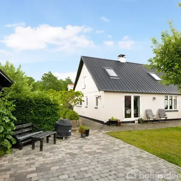 Et hvidt hus med sort tag omkranses af en flisebelagt terrasse med havemøbler og grill. En grøn have med træer omgiver huset under en skyfri blå himmel.